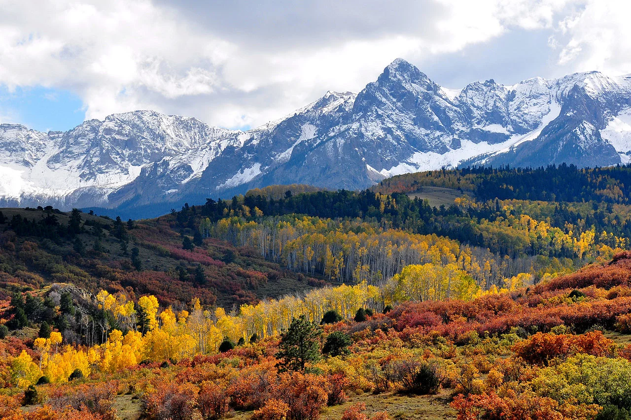 Colorado mountainside blanketed in gold, orange, and red aspen groves beneath snow-dusted peaks in fall
